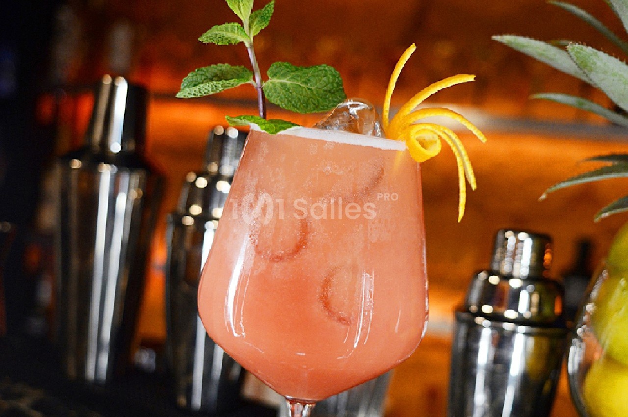Verre de cocktail rose garni de menthe et zeste d'orange, posé sur un bar avec des shakers en arrière-plan.