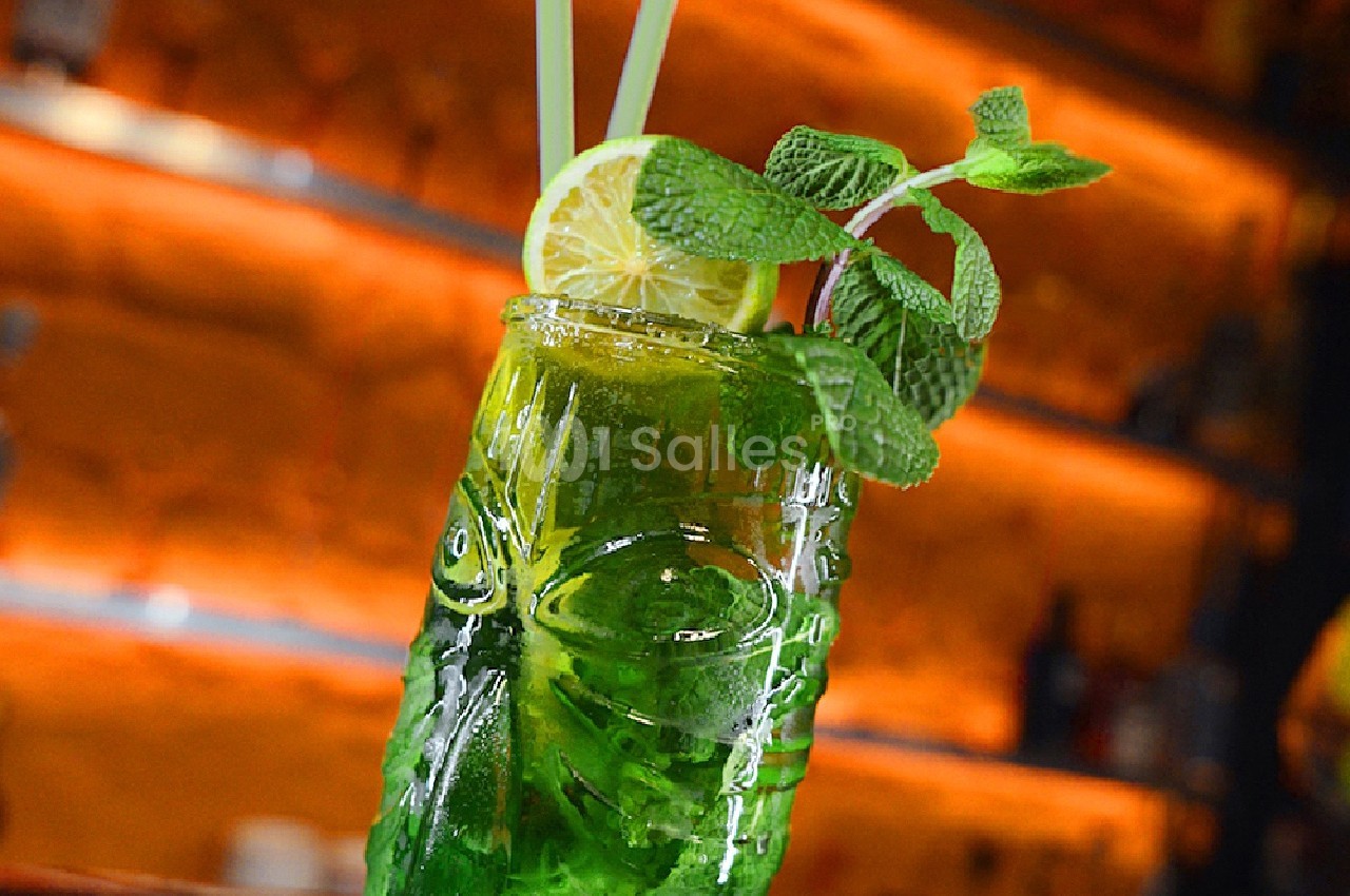 Verre de cocktail vert décoré de menthe fraîche, d'une rondelle de citron et de deux pailles, posé sur un bar.