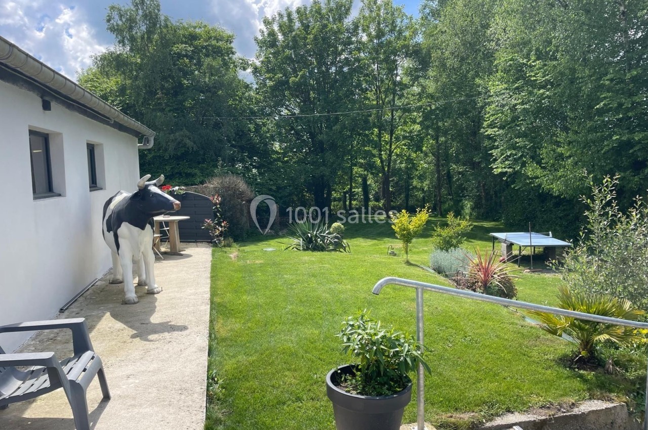 Jardin verdoyant avec pelouse, plantes, trampoline, statue de vache près d'une maison blanche et ciel partiellement nuageux.