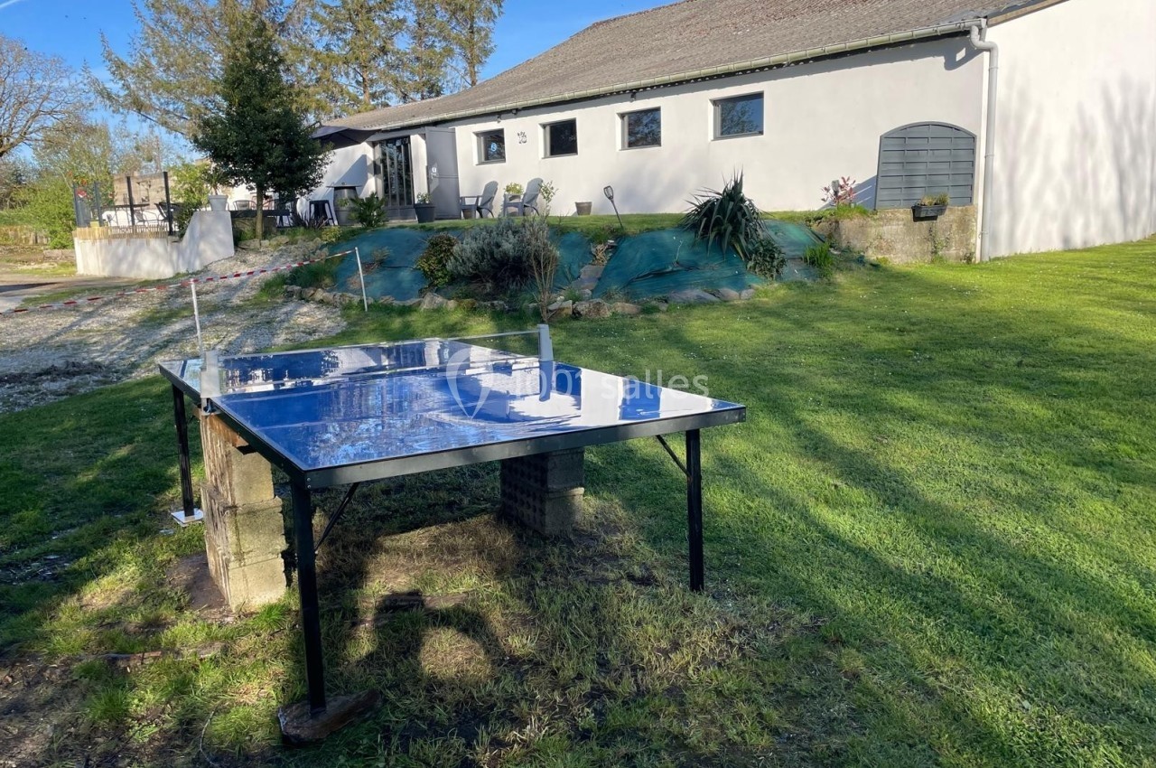 Table de ping-pong extérieure sur une pelouse, devant une maison blanche avec jardin et arbres en arrière-plan.