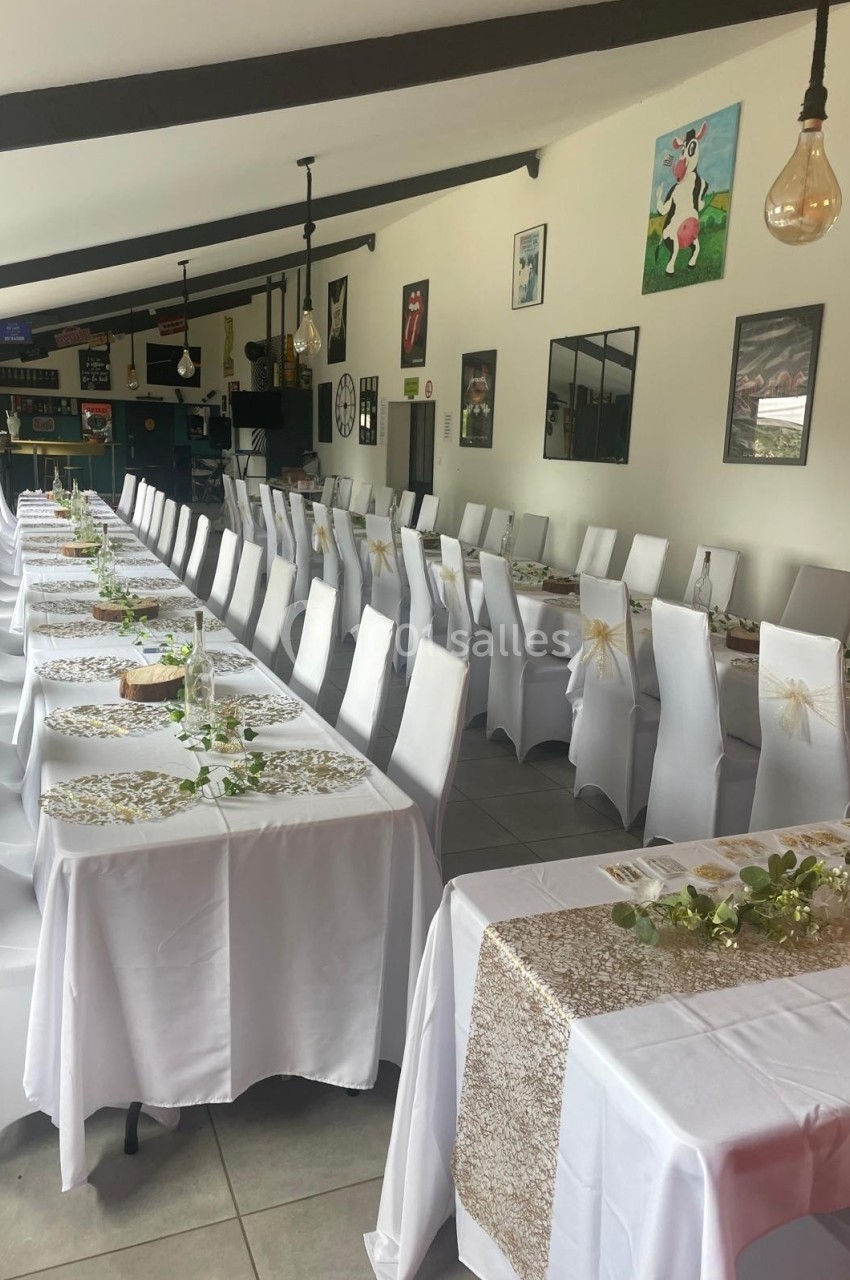 Salle décorée pour un événement avec de longues tables alignées, nappes blanches et chemins de table dorés.