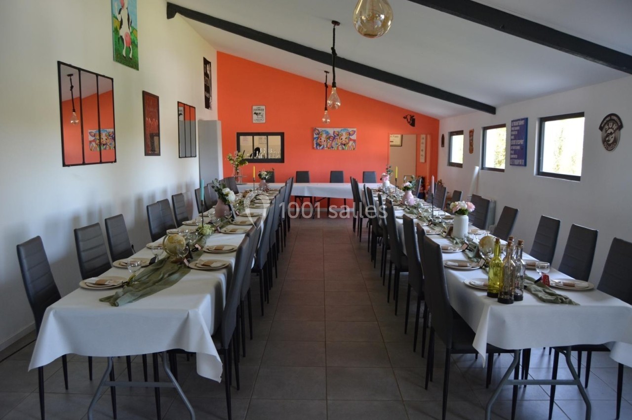 Salle décorée avec deux longues tables dressées pour un repas, entourées de chaises, avec un mur orange en arrière-plan.