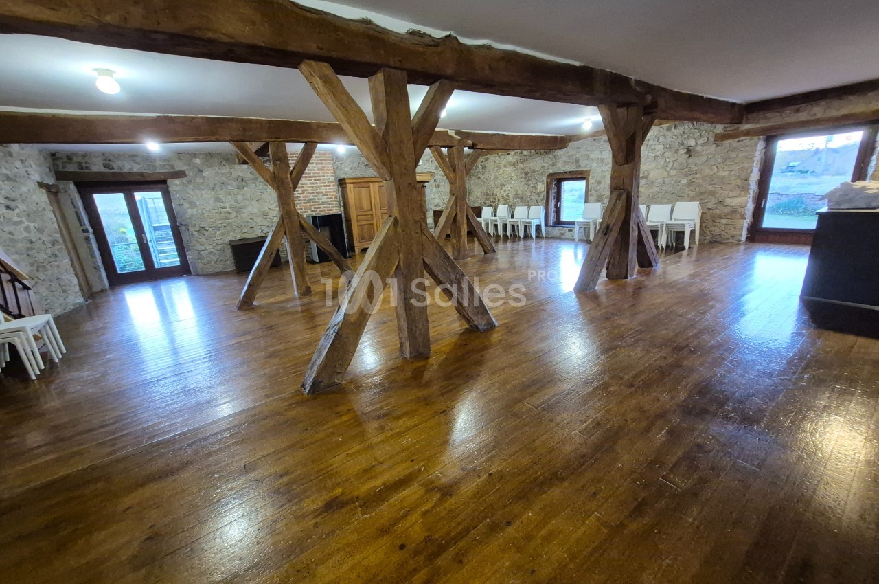Salle spacieuse avec parquet en bois, poutres apparentes et murs en pierre, éclairée par des fenêtres et des luminaires.