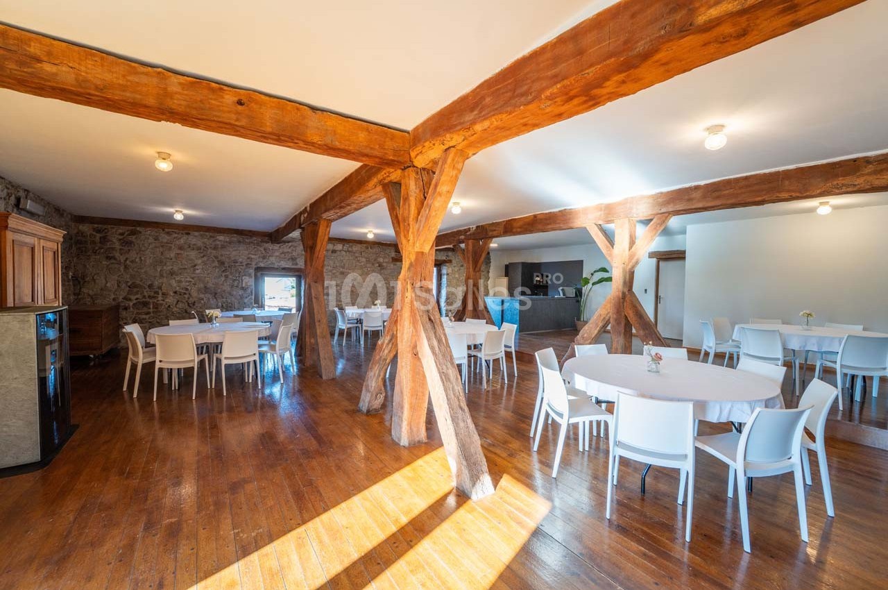 Salle lumineuse avec poutres en bois apparentes, tables rondes blanches et chaises, sur un sol en parquet.