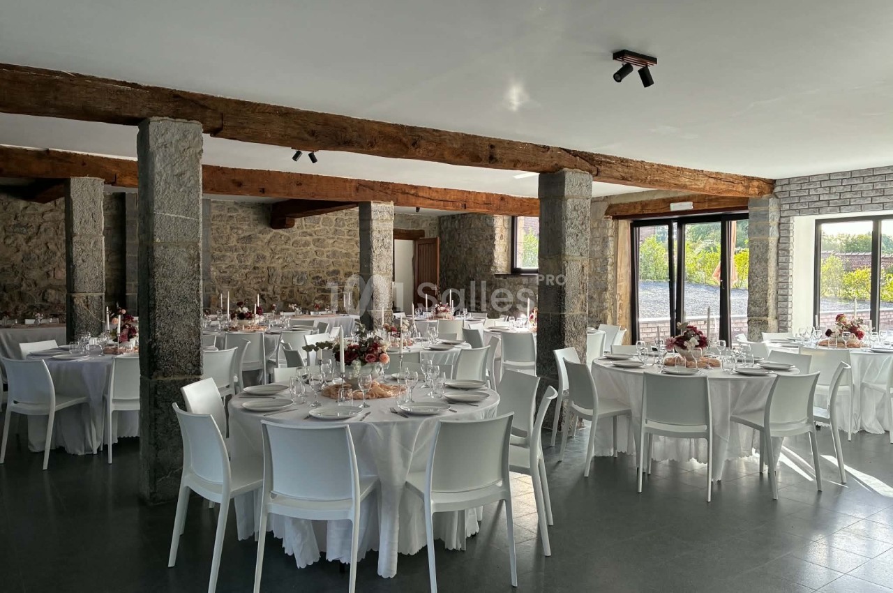 Salle de réception lumineuse avec des tables rondes dressées, chaises blanches et poutres apparentes en bois.