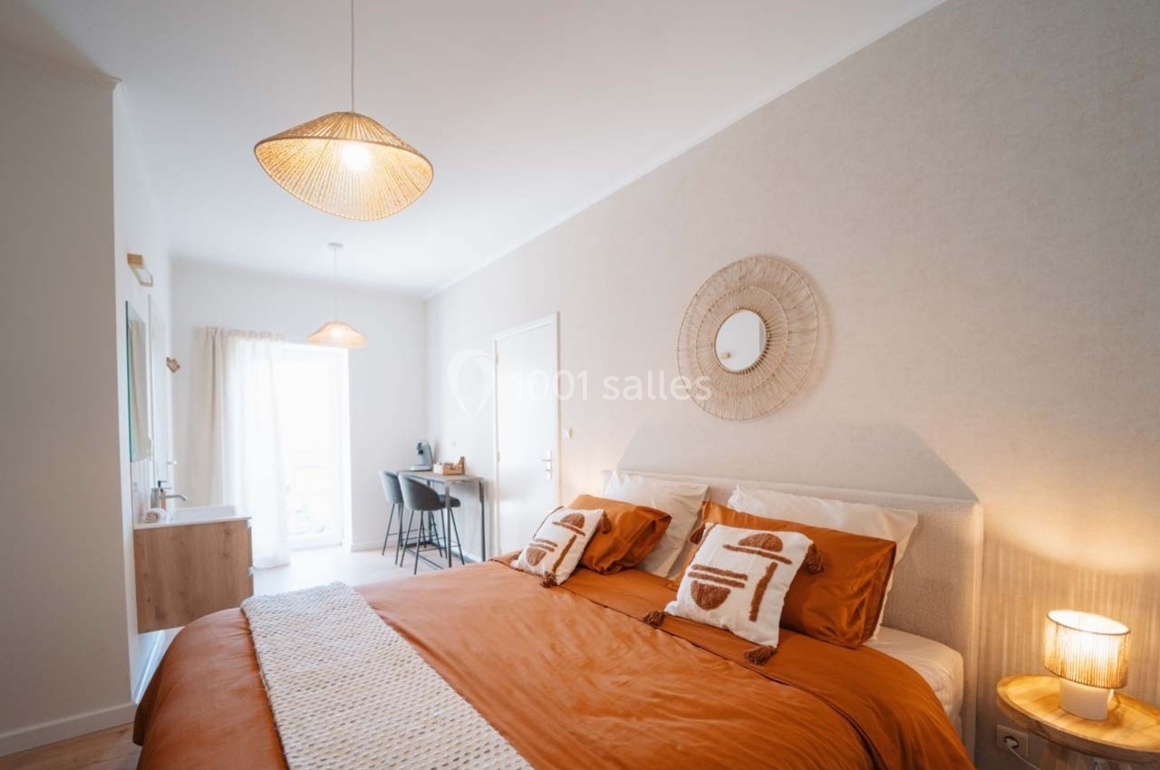 Chambre lumineuse avec lit double, linge orange, bureau, miroir mural et luminaires en rotin.