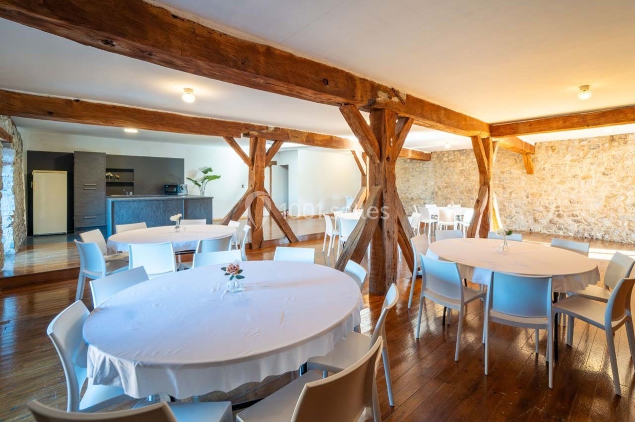 Salle lumineuse avec poutres en bois, murs en pierre et tables rondes dressées avec nappes blanches.