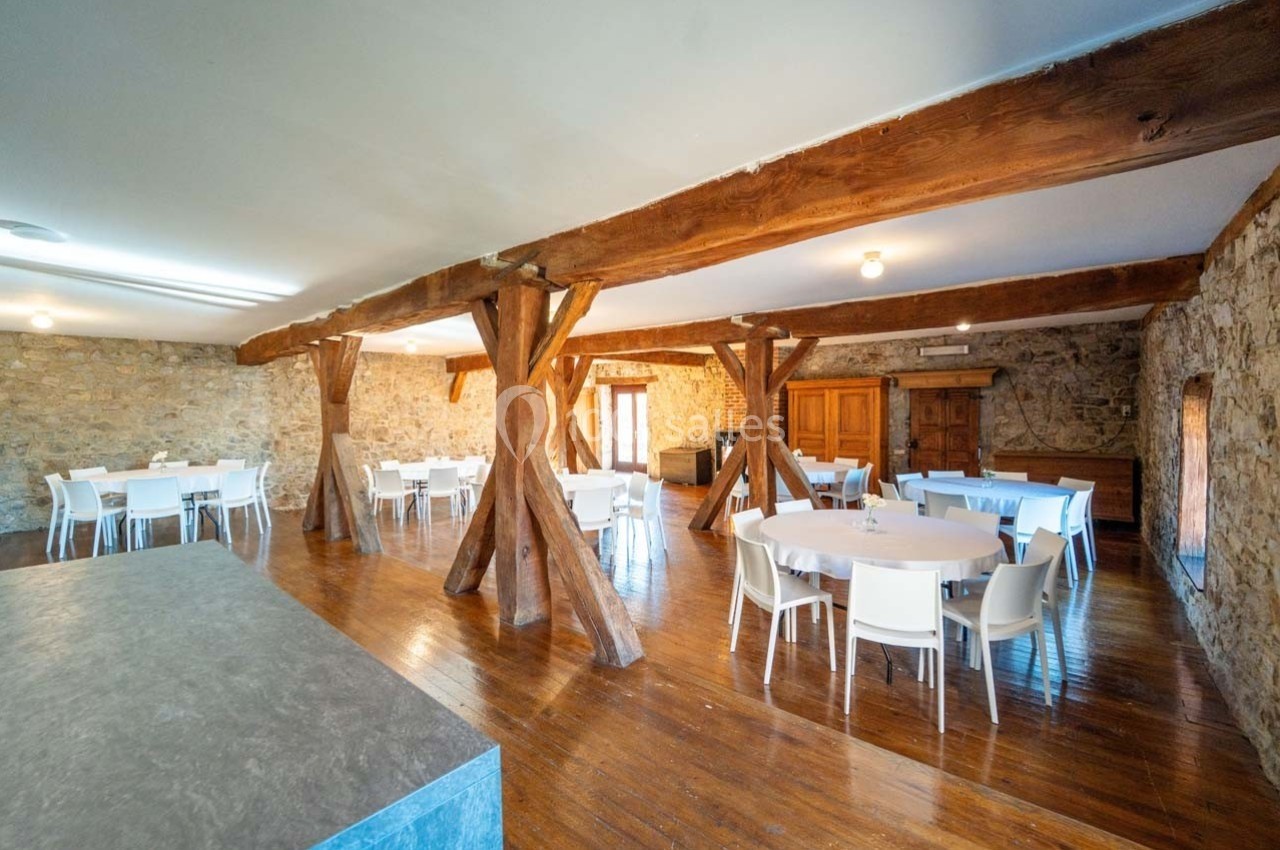 Salle avec murs en pierre, poutres en bois apparentes, tables rondes et chaises blanches disposées sur un sol en parquet.