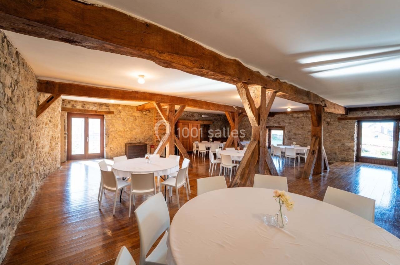 Salle avec murs en pierre, poutres en bois apparentes, tables rondes dressées et lumière naturelle.