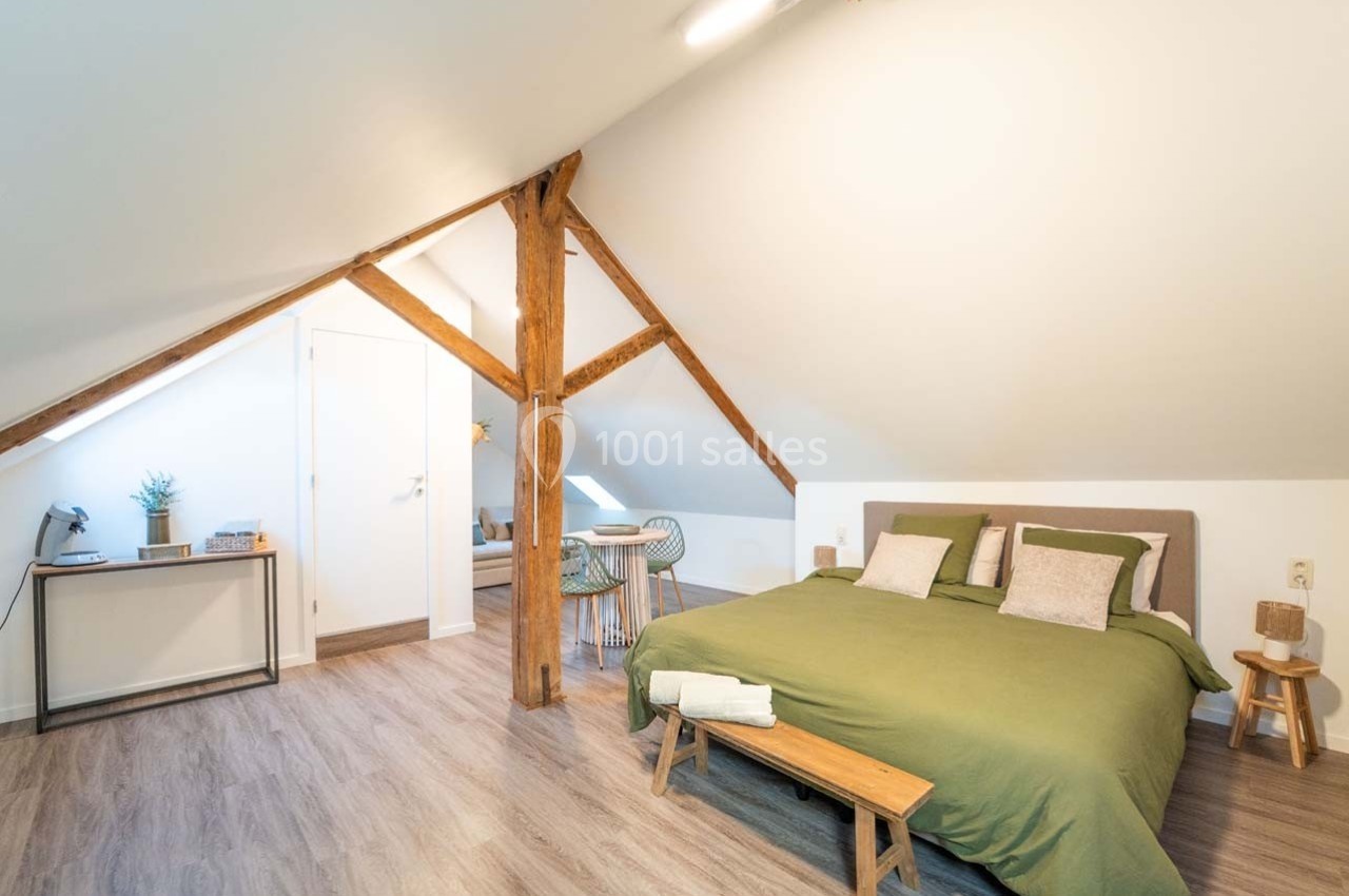 Chambre mansardée lumineuse avec lit double, poutres apparentes, bureau, et coin repas en arrière-plan.