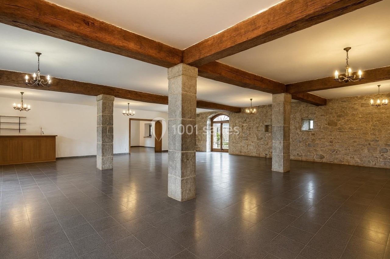 Salle spacieuse avec sol en carrelage, murs en pierre, poutres apparentes et plusieurs lustres suspendus.