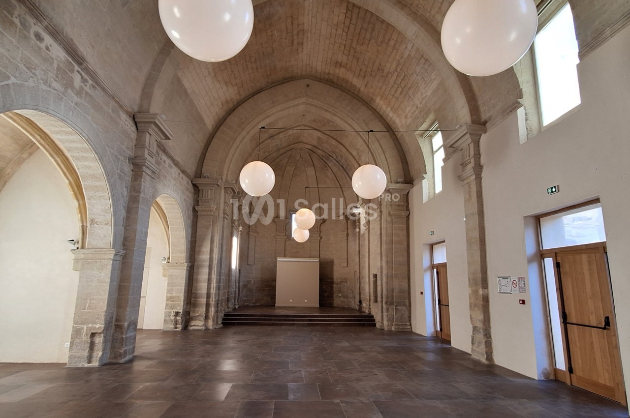 Salle voûtée en pierre avec des arches, des suspensions lumineuses rondes et des portes en bois.
