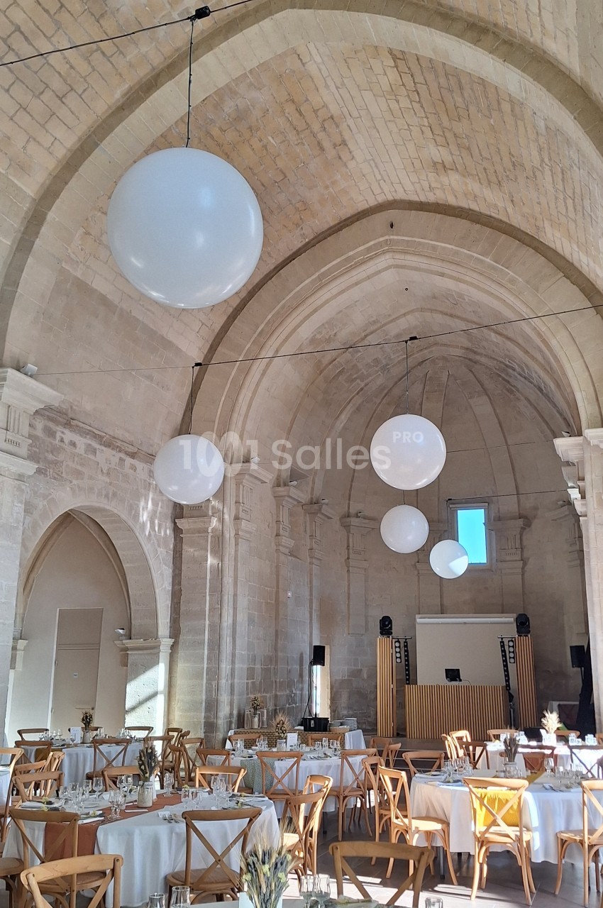 Salle voûtée lumineuse décorée pour un événement, avec tables dressées et grandes suspensions rondes blanches.