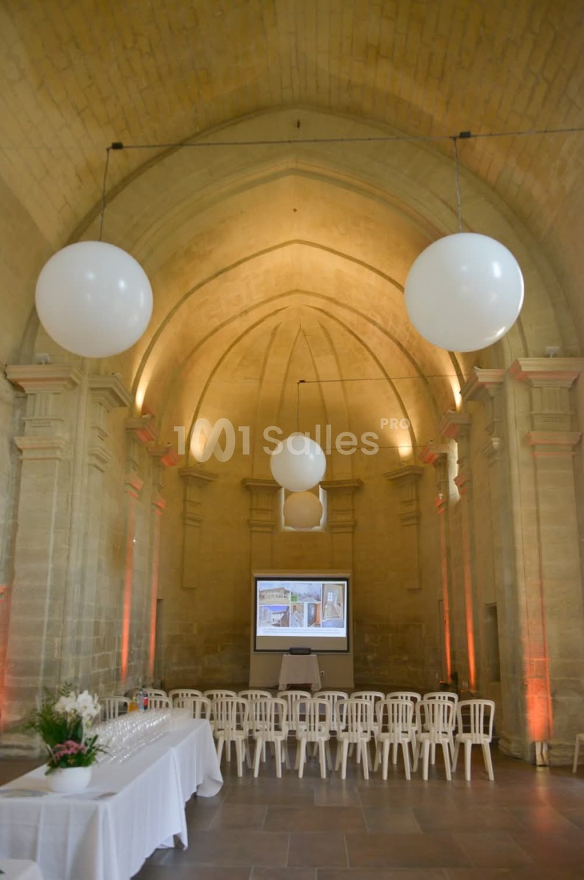 Salle voûtée en pierre avec des chaises alignées, écran de projection, tables dressées et décorations lumineuses.
