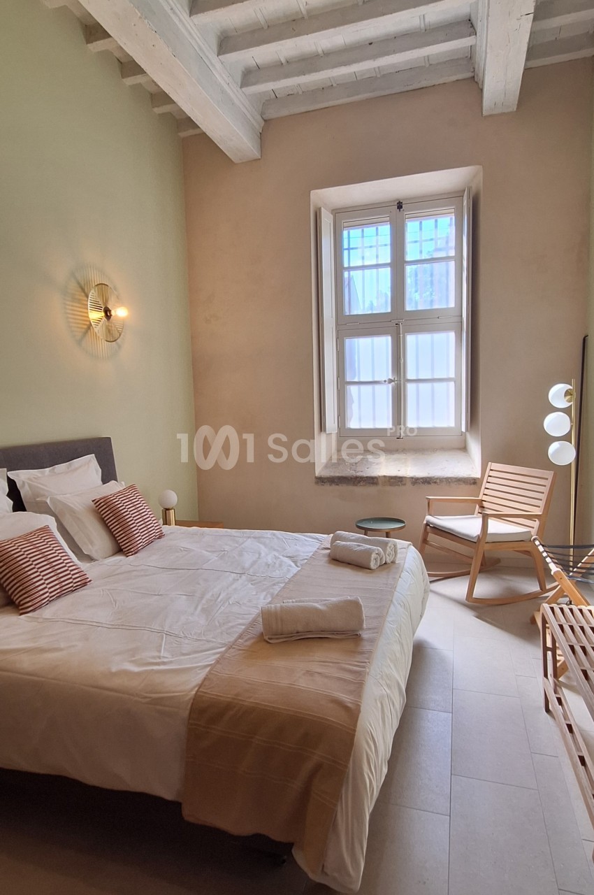 Chambre lumineuse avec lit double, murs clairs, plafond en poutres apparentes, chaise en bois et fenêtre à petits carreaux.