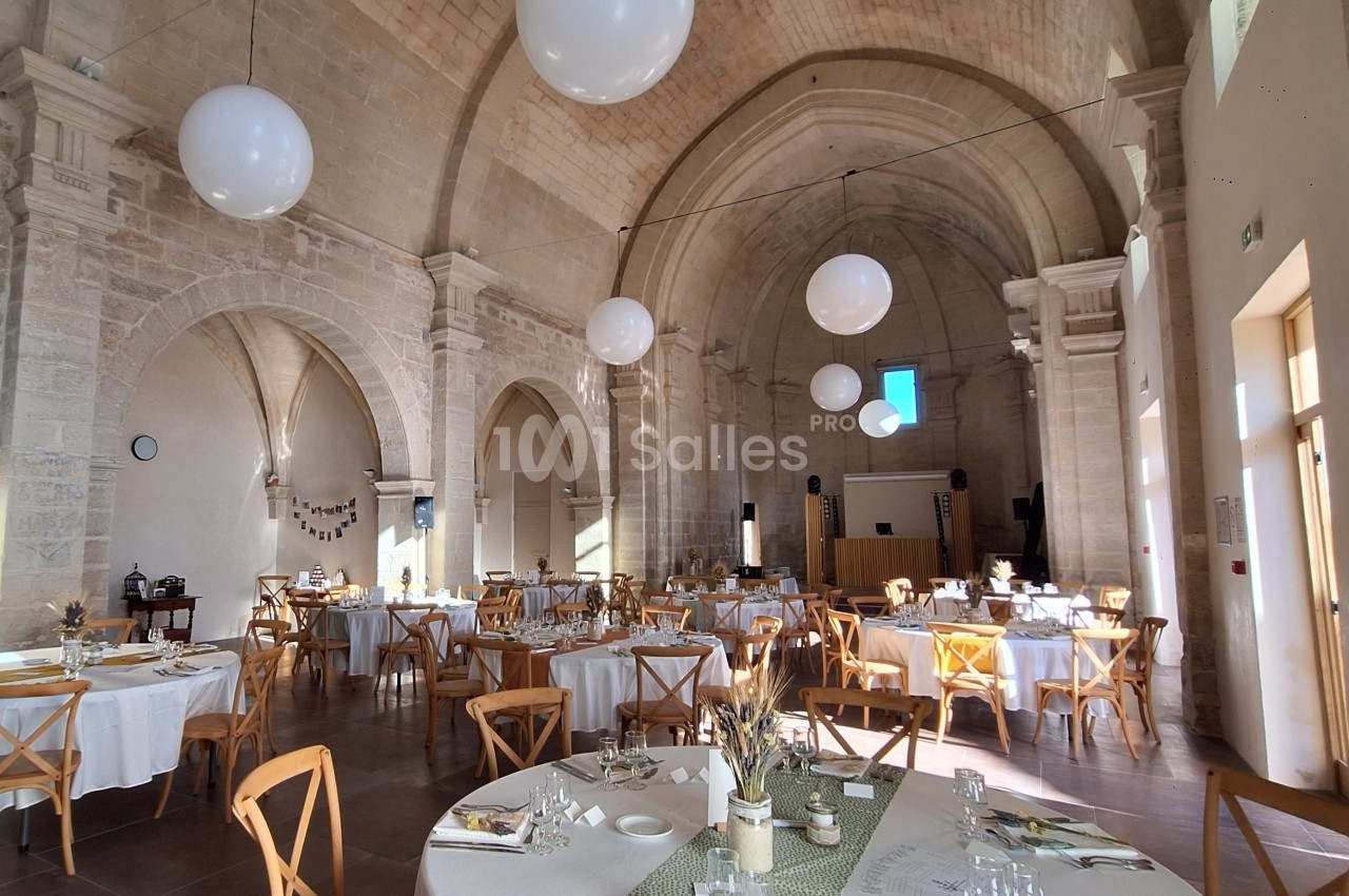 Salle voûtée en pierre aménagée pour un repas, avec tables dressées, chaises en bois et décorations lumineuses.