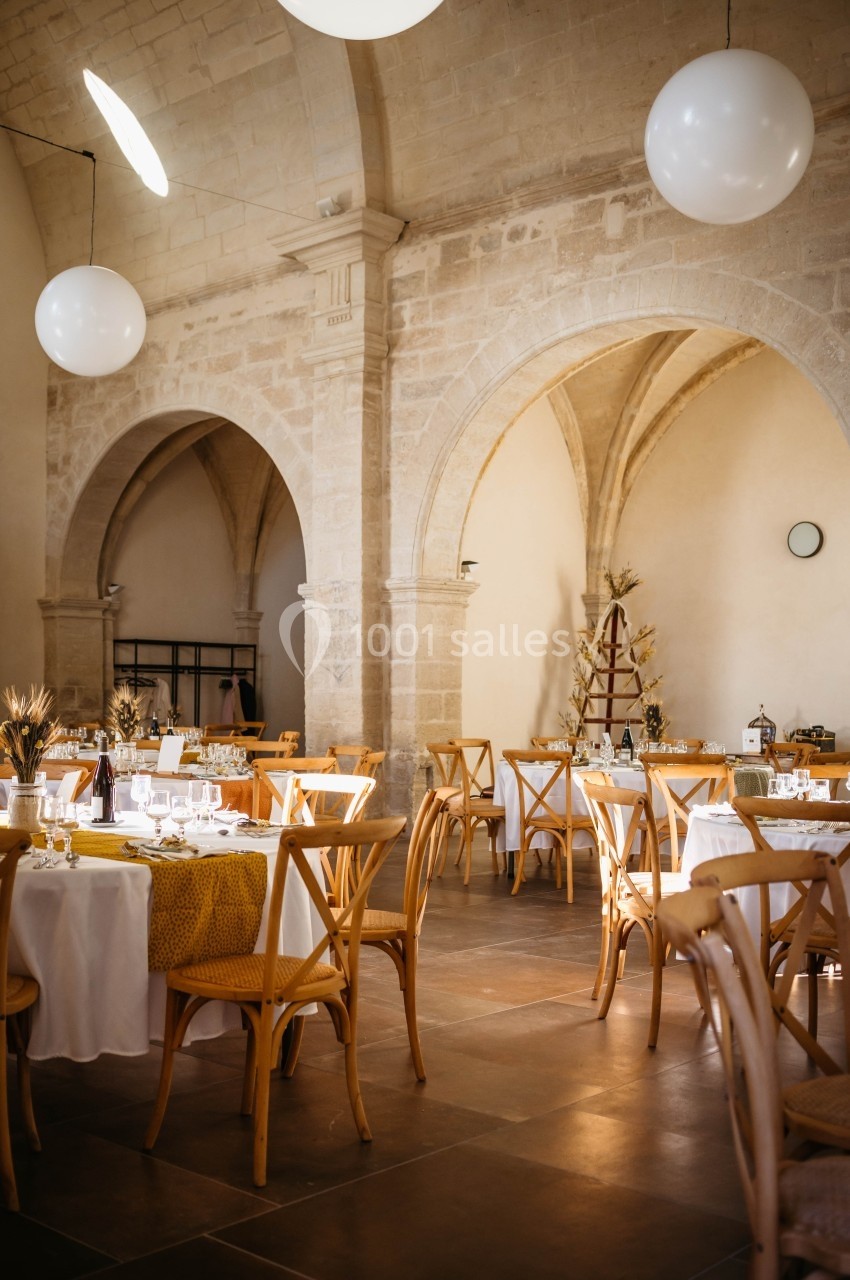 Salle de réception lumineuse avec tables rondes dressées, chaises en bois et murs en pierre voûtés.