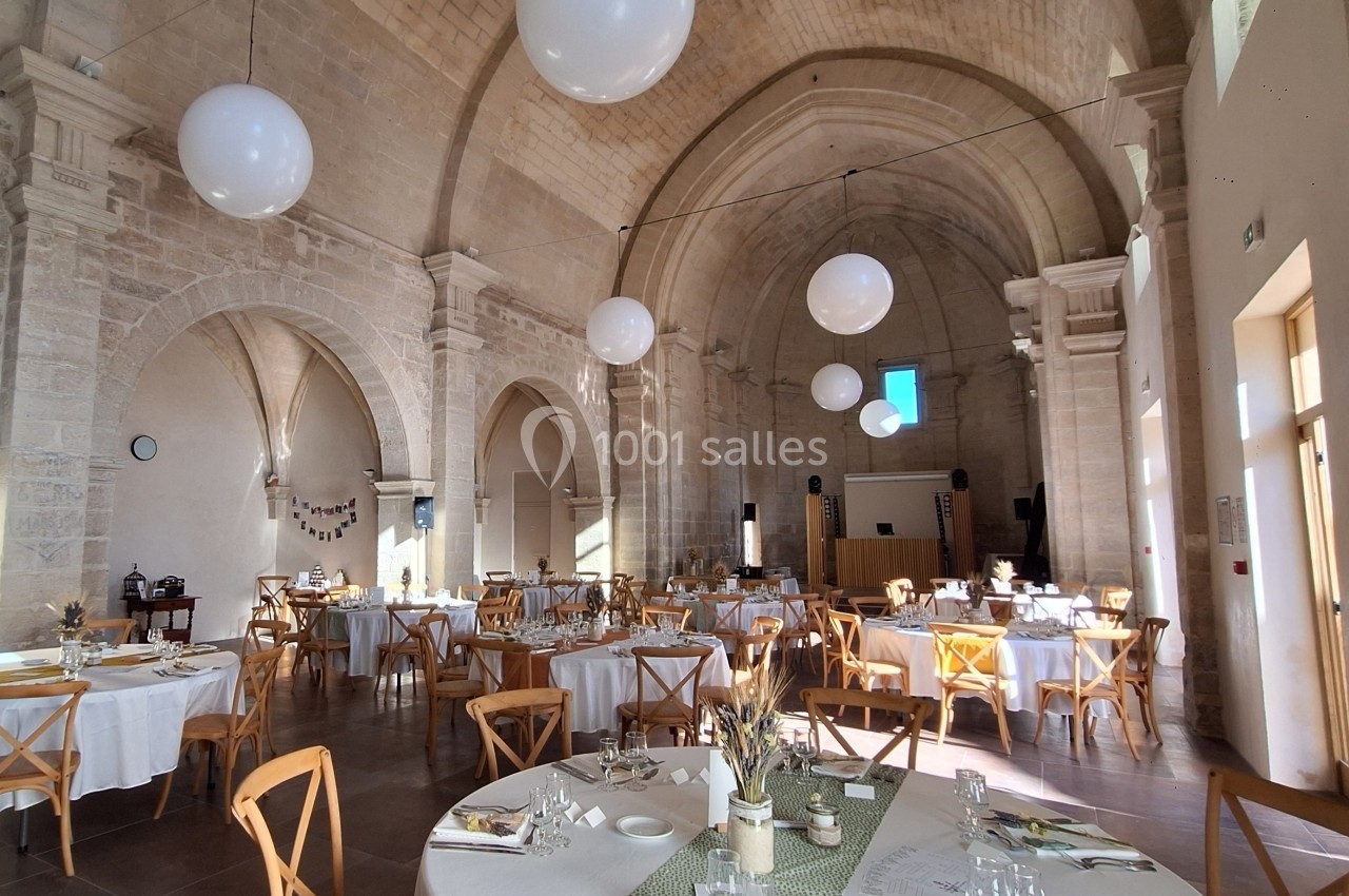 Salle voûtée en pierre aménagée pour un repas, avec tables dressées, chaises en bois et décorations lumineuses.