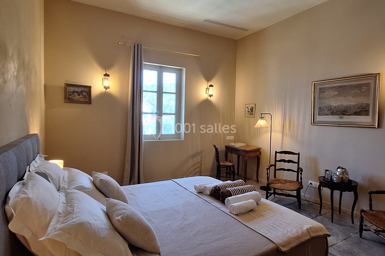 Chambre lumineuse avec lit double, draps blancs, murs beige, bureau en bois, chaises anciennes et décoration murale.