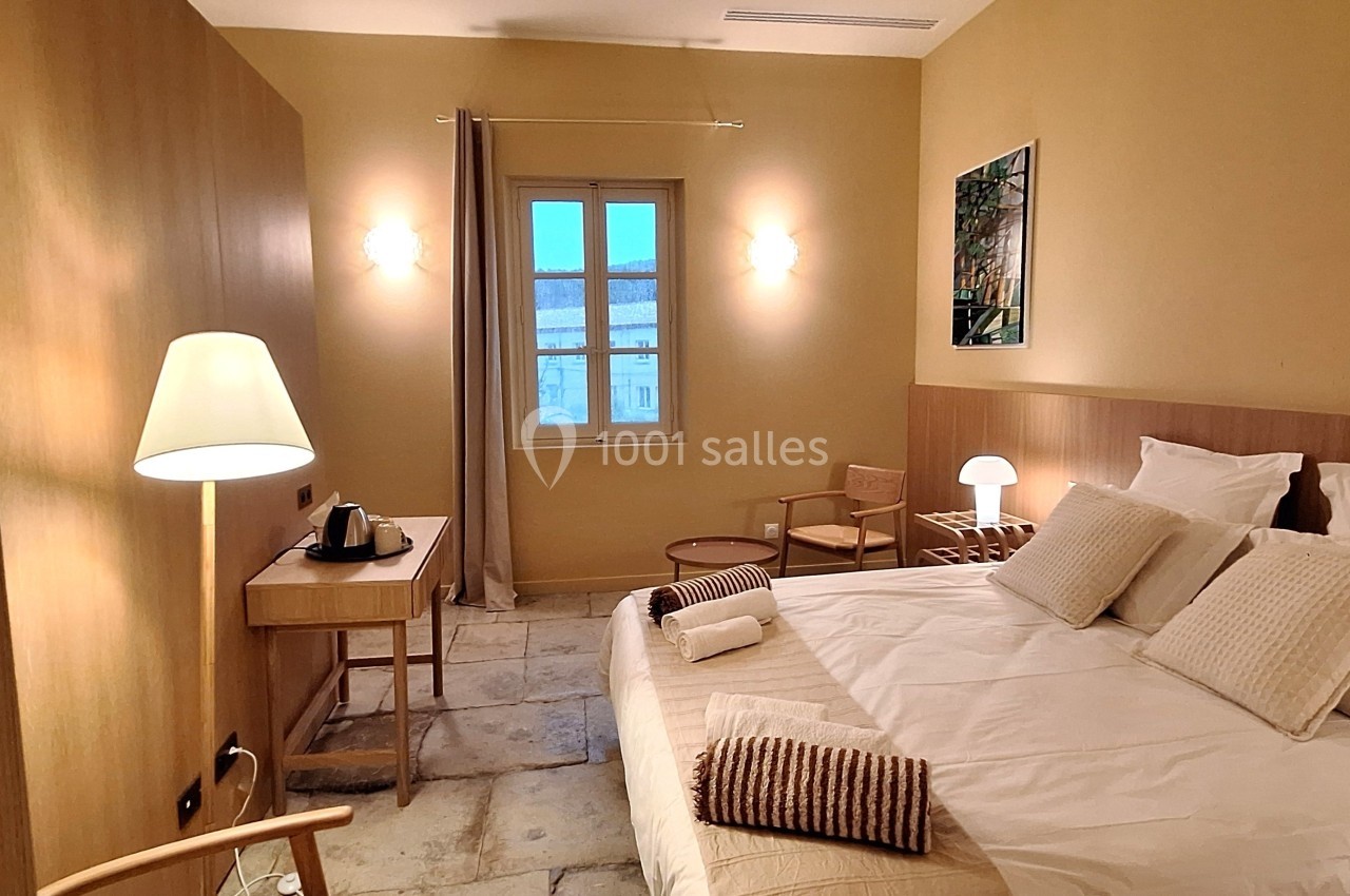 Chambre lumineuse avec lit double, bureau, fauteuils en bois, lampes et fenêtre donnant sur un paysage extérieur.