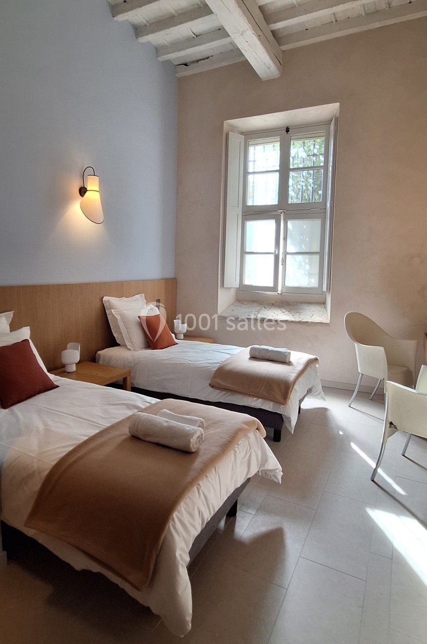 Deux lits simples avec linge beige et coussins rouges dans une chambre lumineuse avec fenêtre et plafond en poutres…