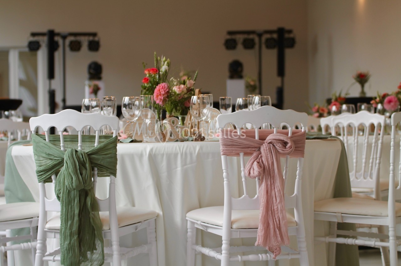 Salle décorée pour un événement avec des chaises blanches ornées de nœuds en tissu rose et vert autour d'une table dressée.