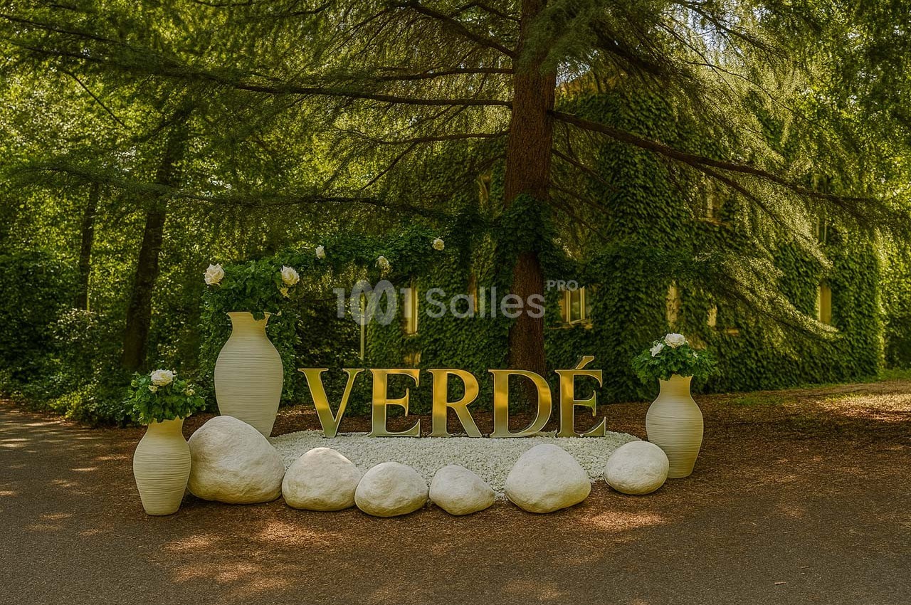 Installation décorative avec des vases blancs, des plantes et des lettres formant ’VERDÉ’, entourée de pierres blanches…