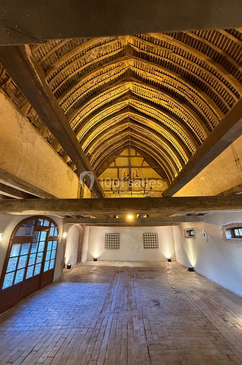 Charpente en bois apparente d'une grande salle avec murs clairs, sol en briques et larges portes vitrées.
