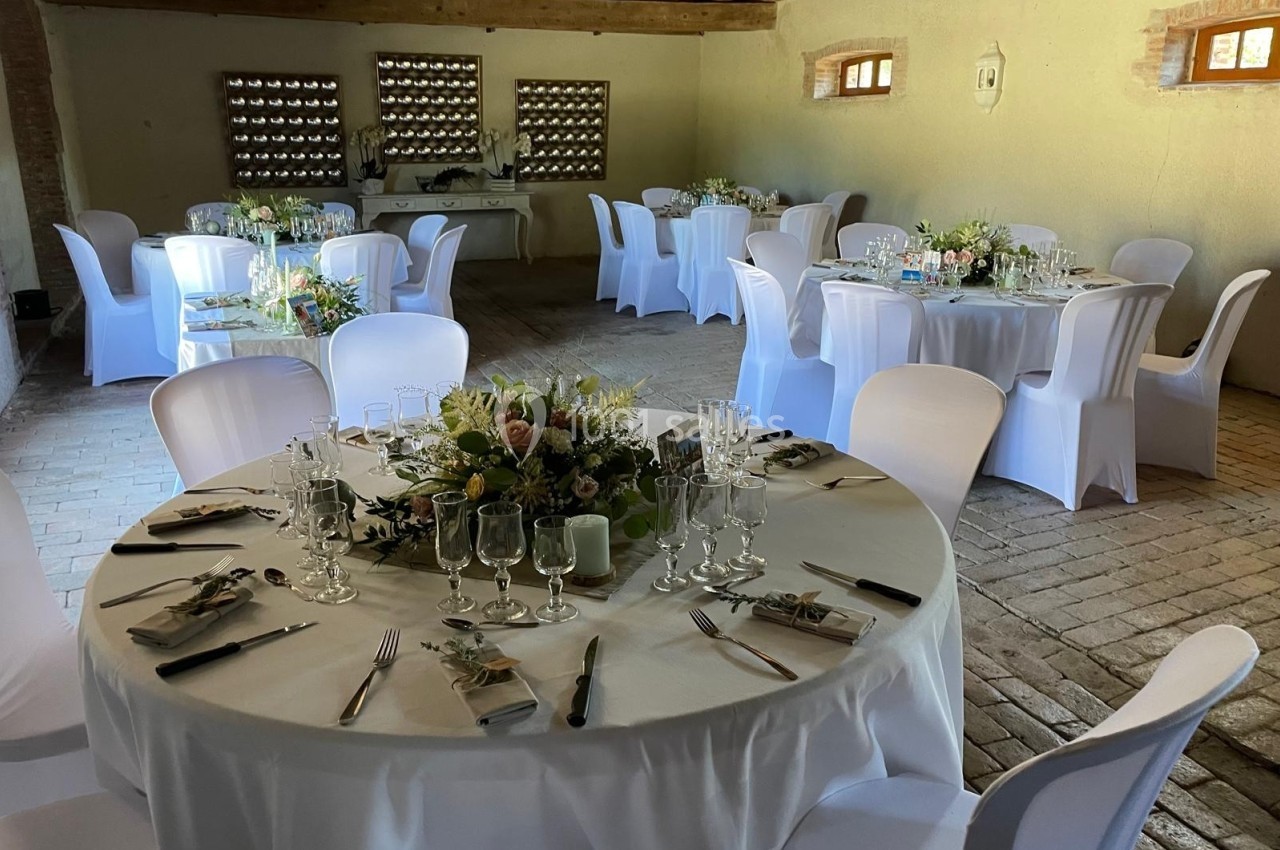 Salle de réception avec tables rondes dressées, nappes blanches, chaises houssées et décorations florales.