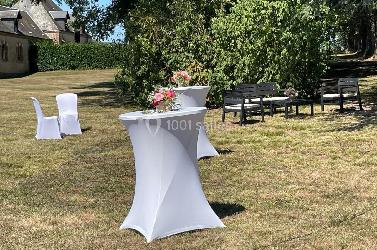 Tables hautes drapées de blanc et décorées de fleurs dans un jardin, avec des chaises et des arbres en arrière-plan.