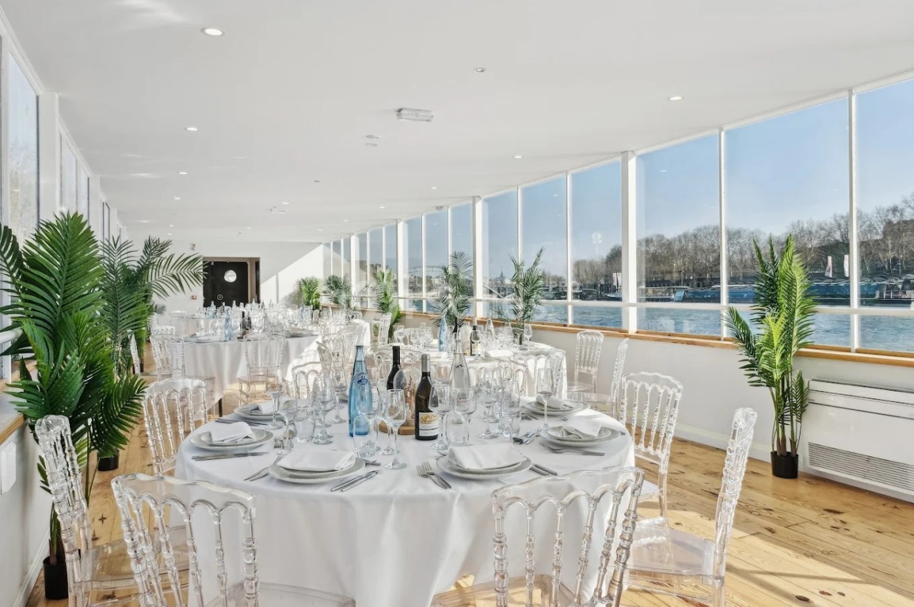 Salle de réception lumineuse avec tables rondes dressées, baies vitrées donnant sur une vue fluviale et plantes décoratives.