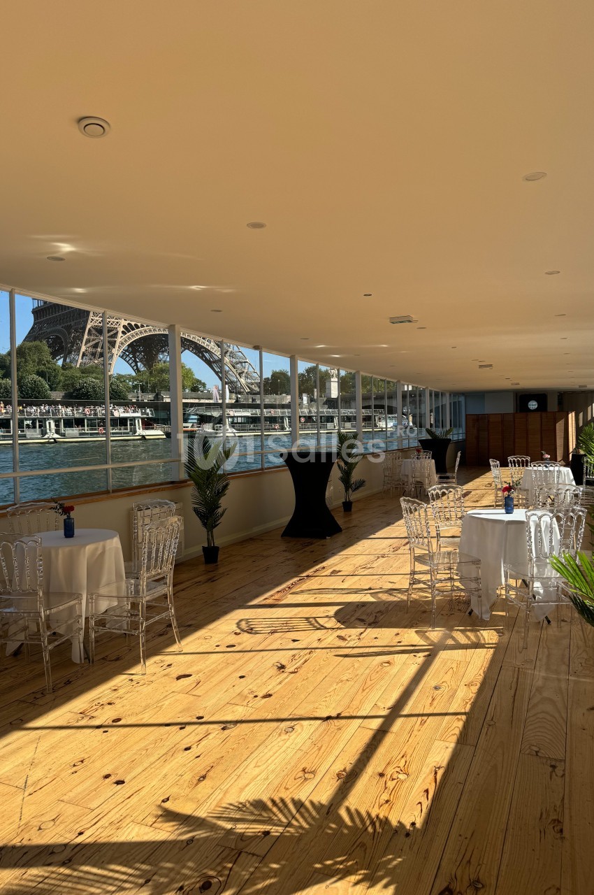 Salle lumineuse avec parquet en bois, tables dressées et vue sur la Tour Eiffel et la Seine à travers de grandes baies…