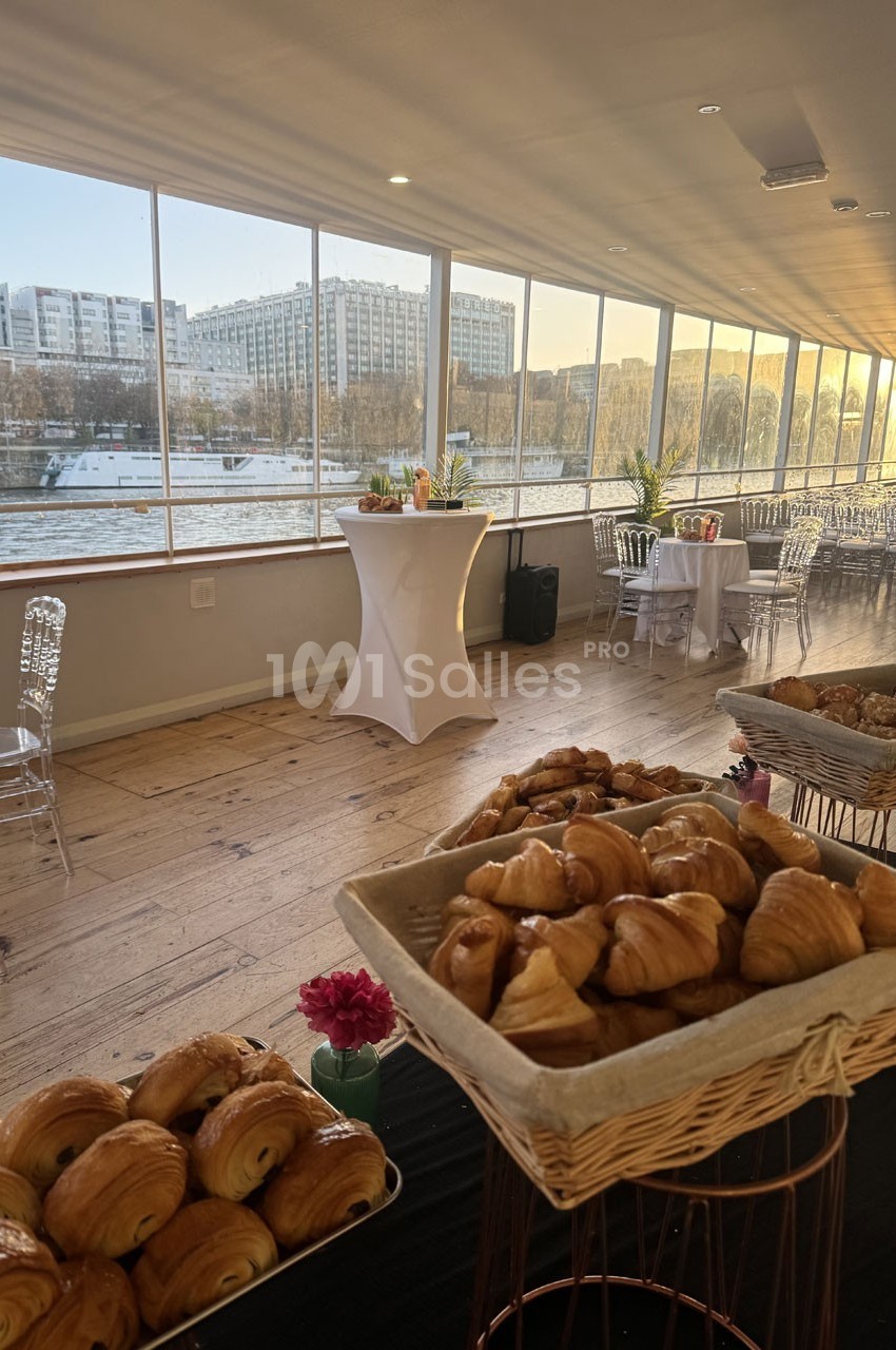 Salle lumineuse avec vue sur la Seine, tables hautes décorées et paniers de viennoiseries au premier plan.