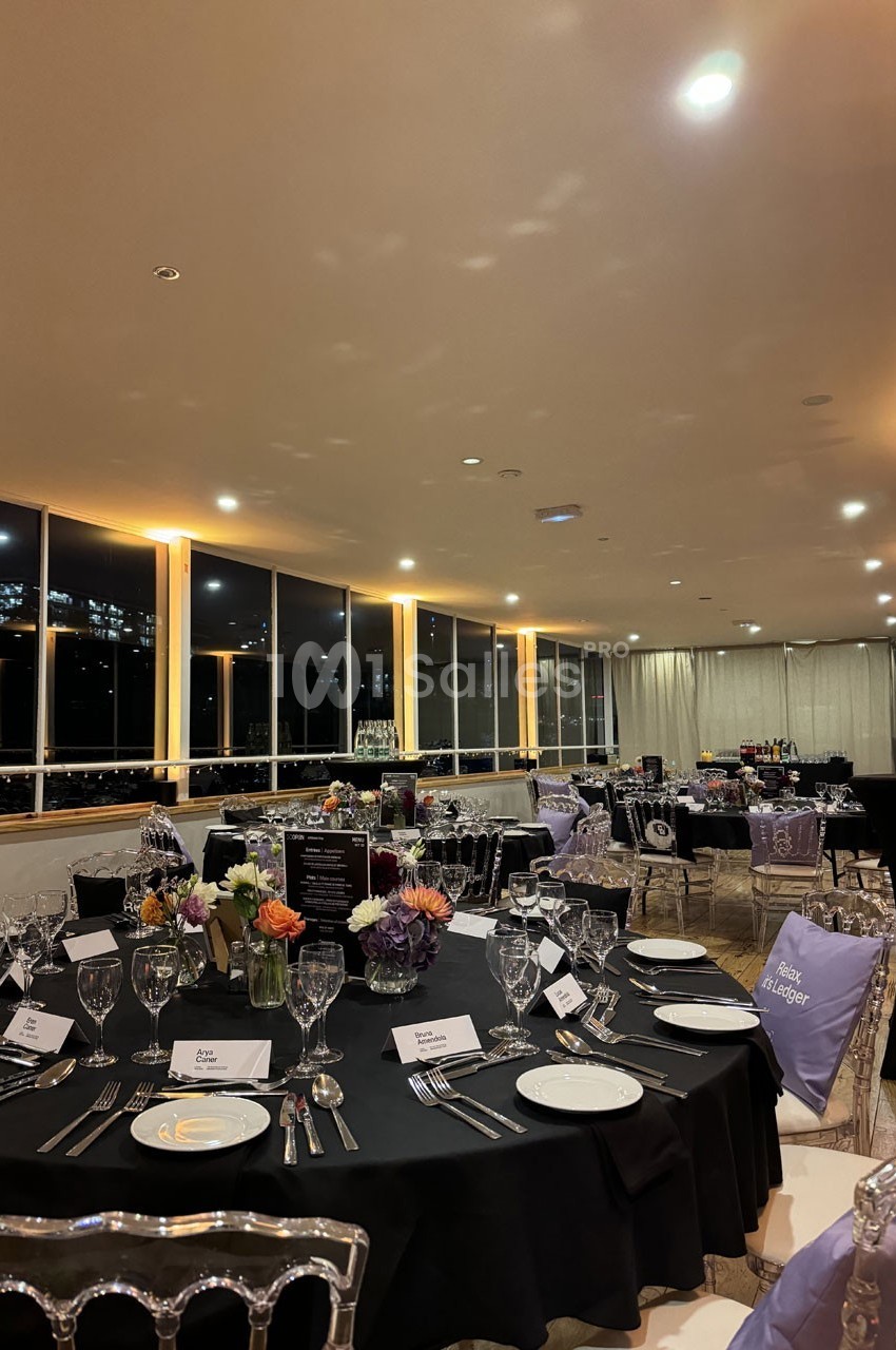 Salle élégamment décorée pour un dîner, avec tables dressées, nappes noires, chaises transparentes et vue nocturne sur la…