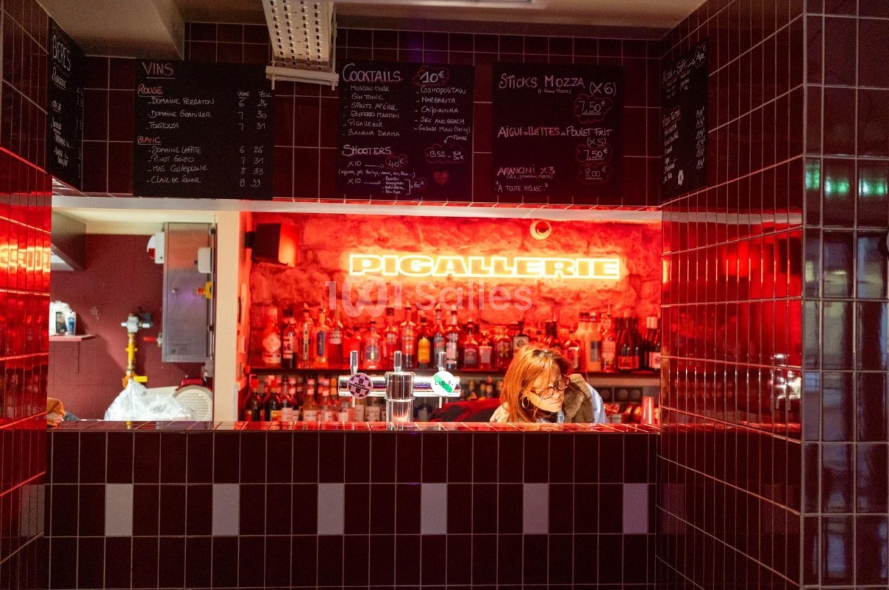 Bar avec comptoir en carrelage rouge, étagères de bouteilles et enseigne lumineuse ’Pigallerie’ en arrière-plan.