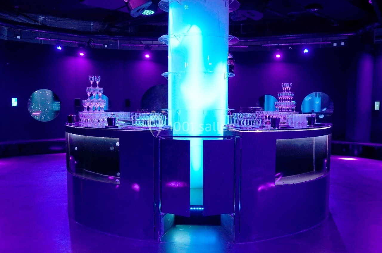 Salle sombre éclairée par des lumières violettes et bleues, avec des tours de verres empilés sur des comptoirs circulaires.