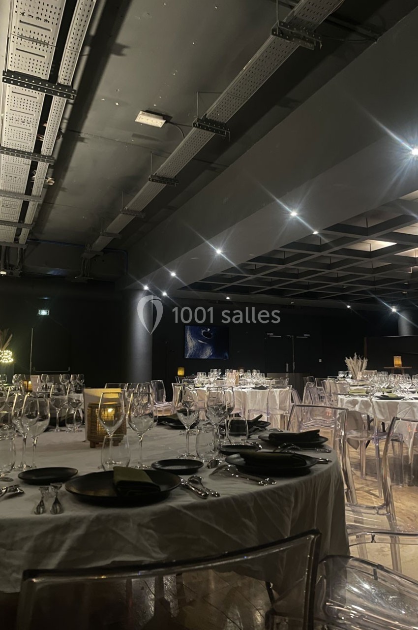Salle de réception sombre avec tables dressées, nappes blanches, vaisselle élégante et chaises transparentes.