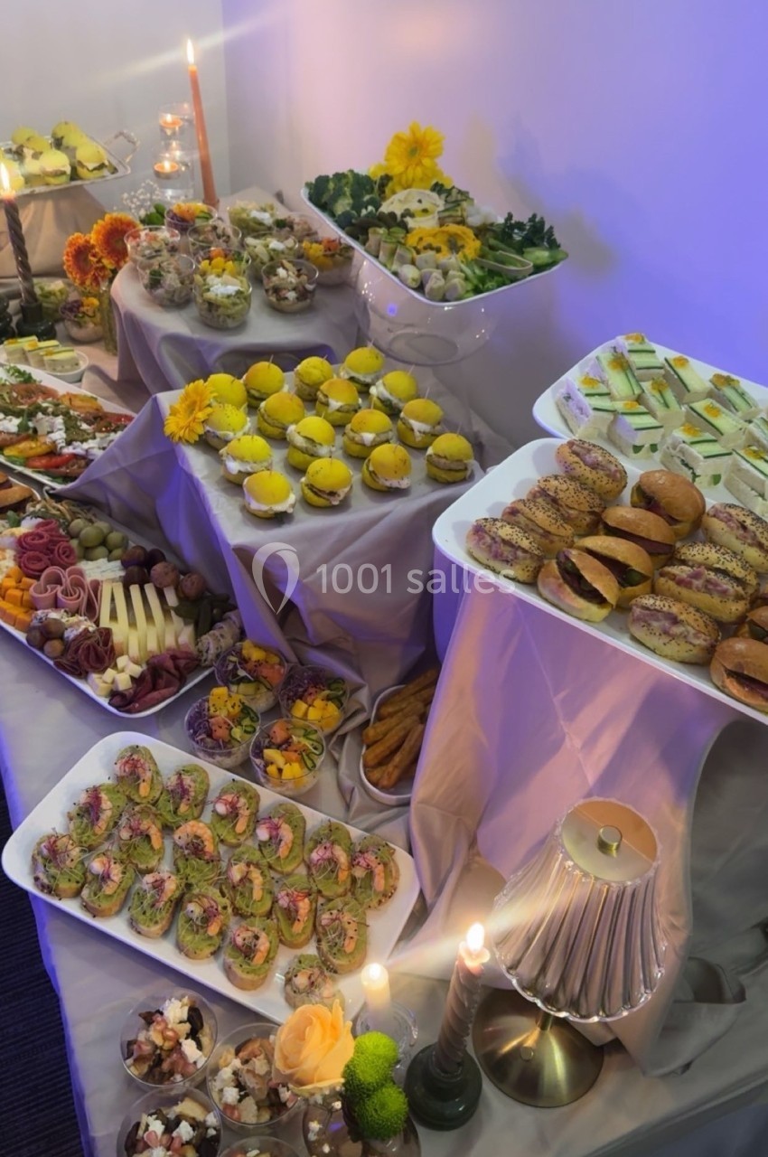 Buffet varié avec amuse-bouches, sandwichs, desserts et décorations florales sur des nappes blanches.