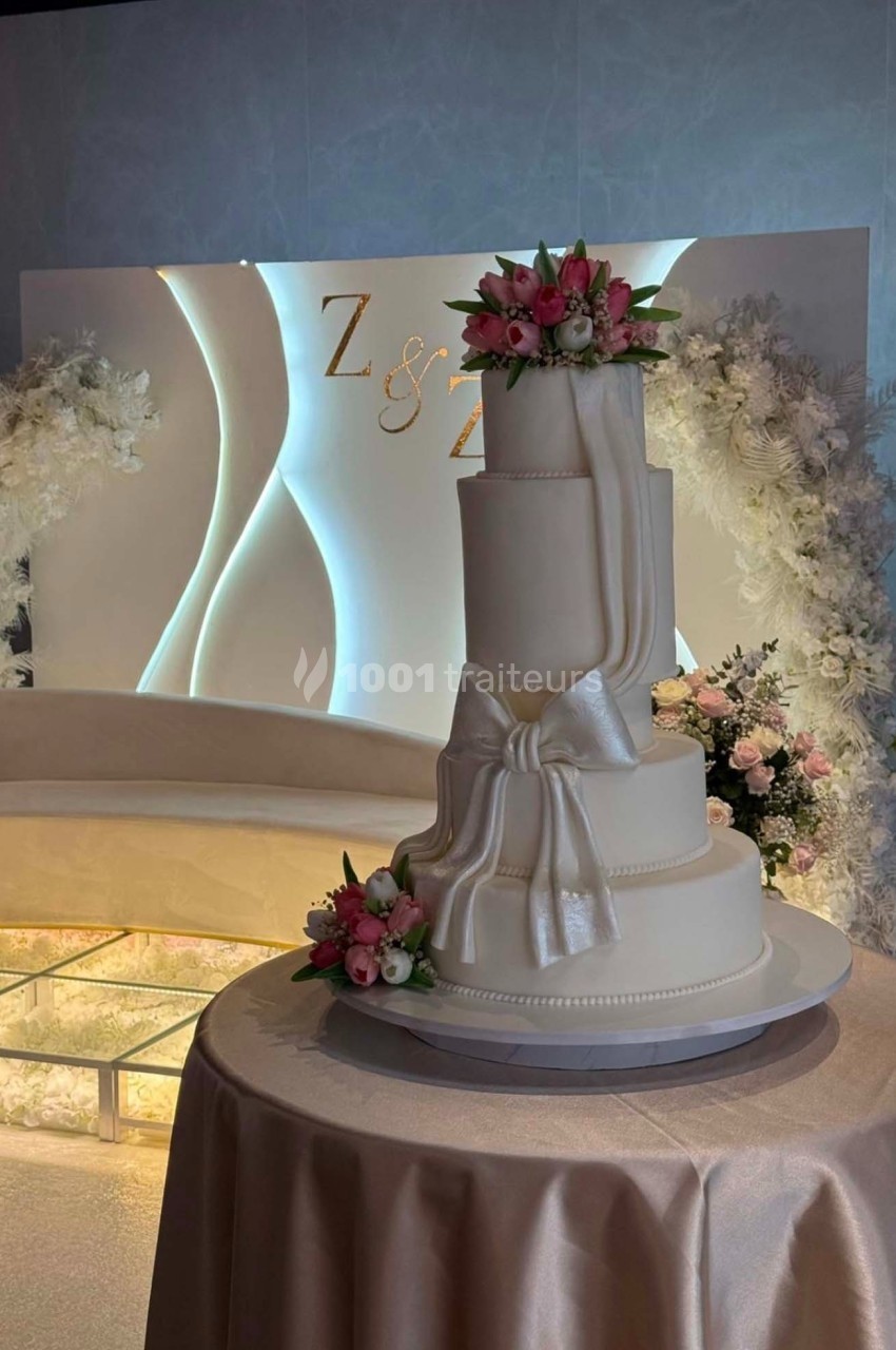 Gâteau de mariage à trois étages décoré de fleurs roses et blanches, avec un nœud en glaçage, sur une table drapée.