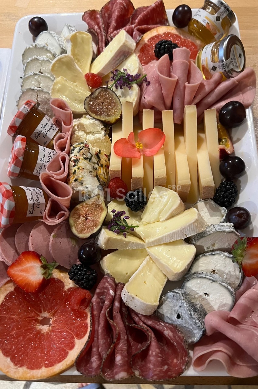 Plateau de charcuteries, fromages variés, fruits frais, confitures et décorations florales sur une table en bois.