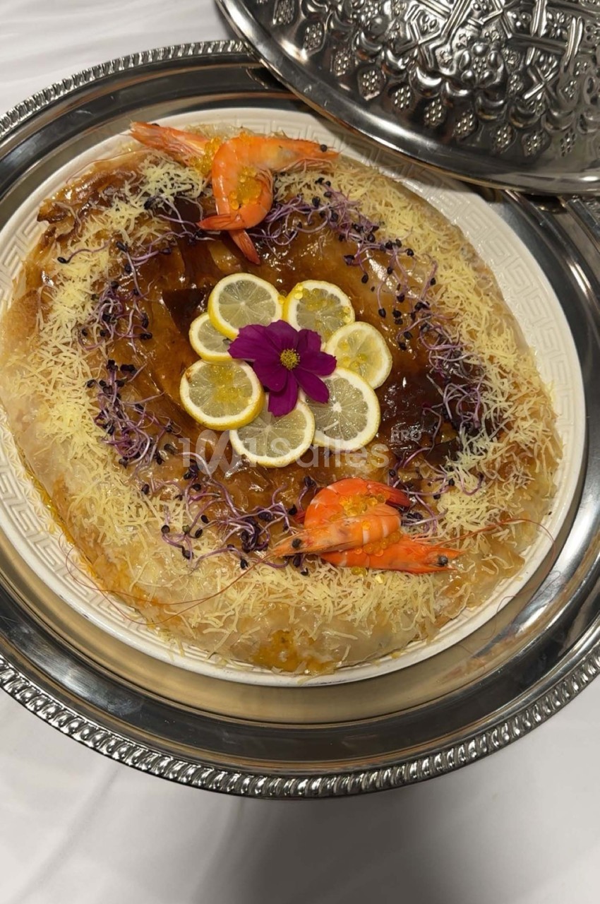 Plat de présentation contenant une pastilla garnie de crevettes, tranches de citron et décorée de fleurs et pousses.