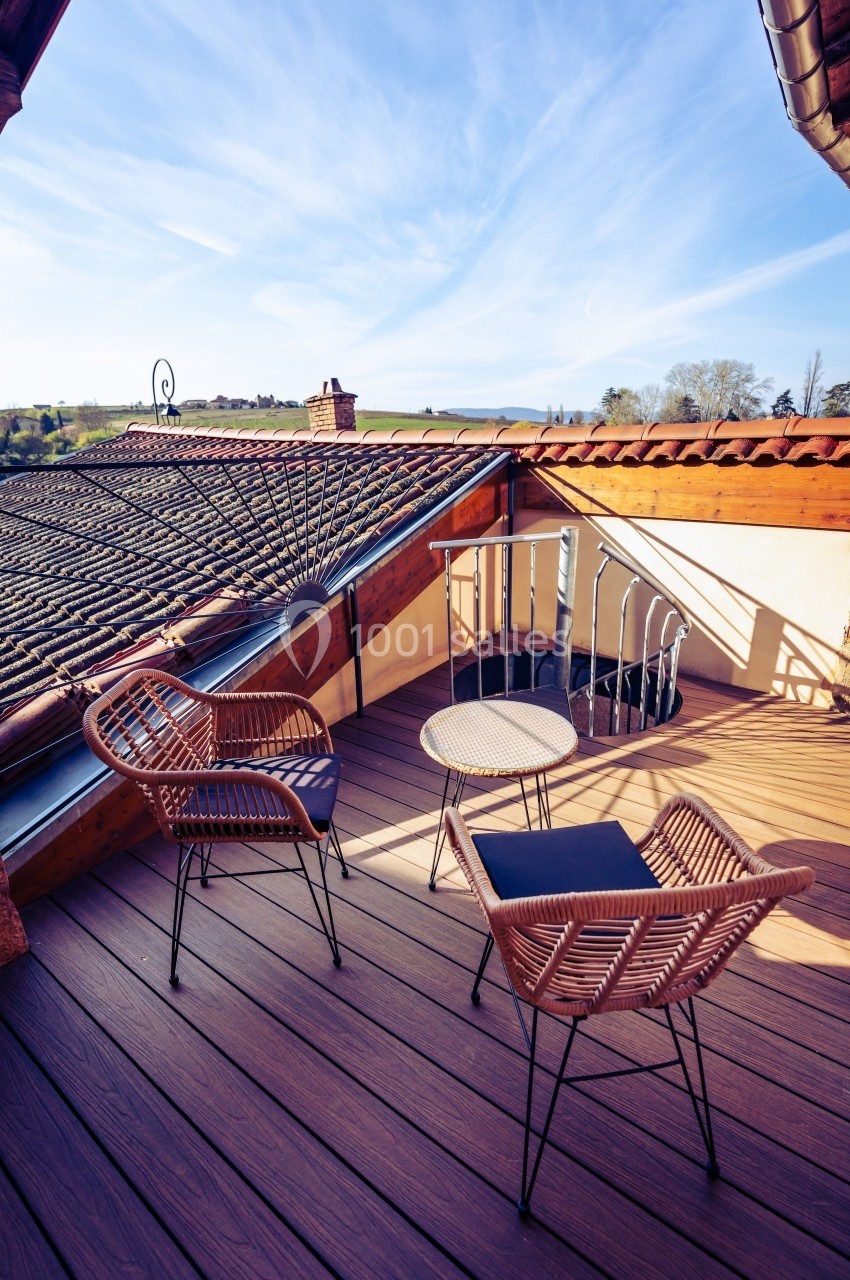 Terrasse en bois avec deux chaises en rotin, une table ronde et vue sur des toits et la campagne environnante.