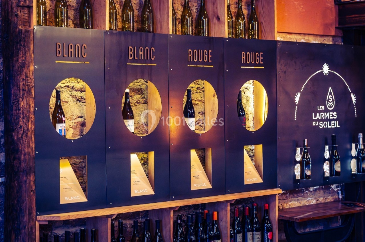 Distributeur de vin en libre-service avec sections pour vin blanc et rouge, entouré de bouteilles sur étagères en bois.