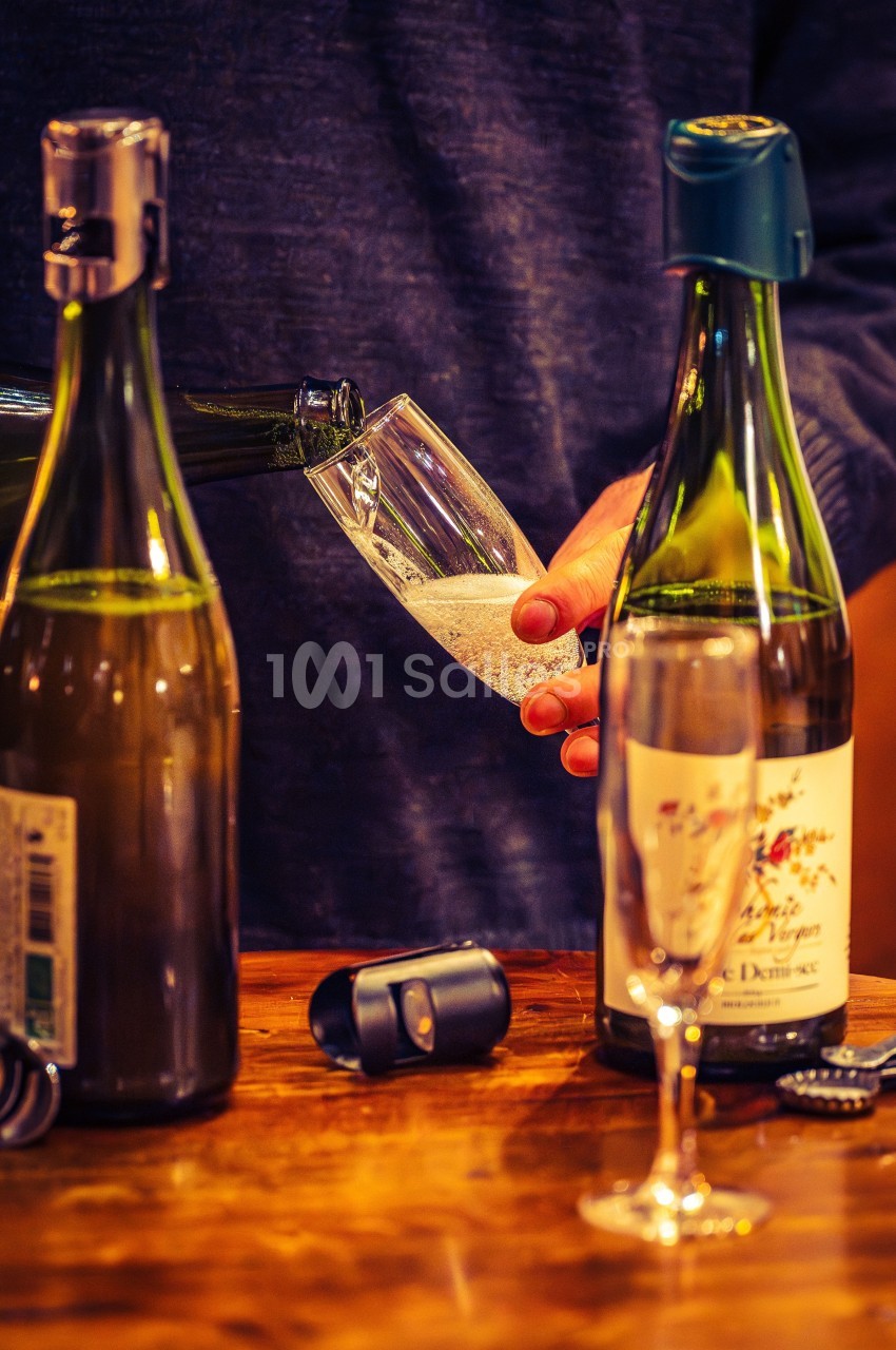 Une personne verse du vin pétillant dans une flûte sur une table en bois, entourée de bouteilles ouvertes.