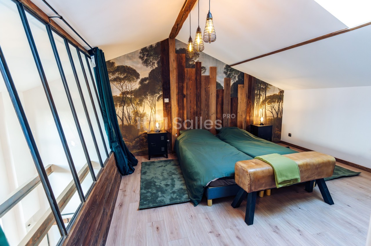 Chambre mansardée avec deux lits simples, mur en bois, papier peint nature, et grandes fenêtres vitrées.