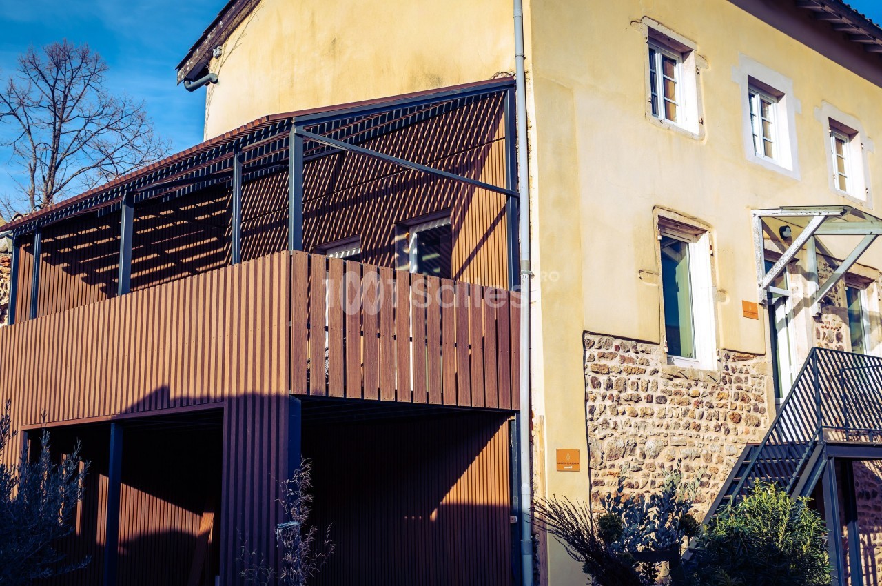 Maison en pierre avec extension en bois, terrasse couverte et escalier extérieur métallique sous un ciel dégagé.