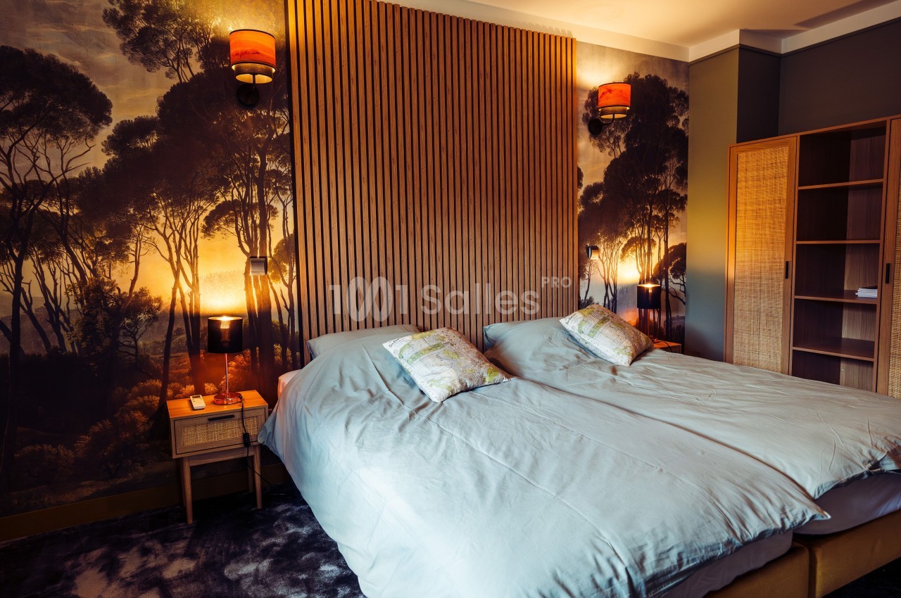 Chambre avec deux lits simples, tête de lit en bois, éclairage chaleureux et mur décoré d'une fresque forestière.