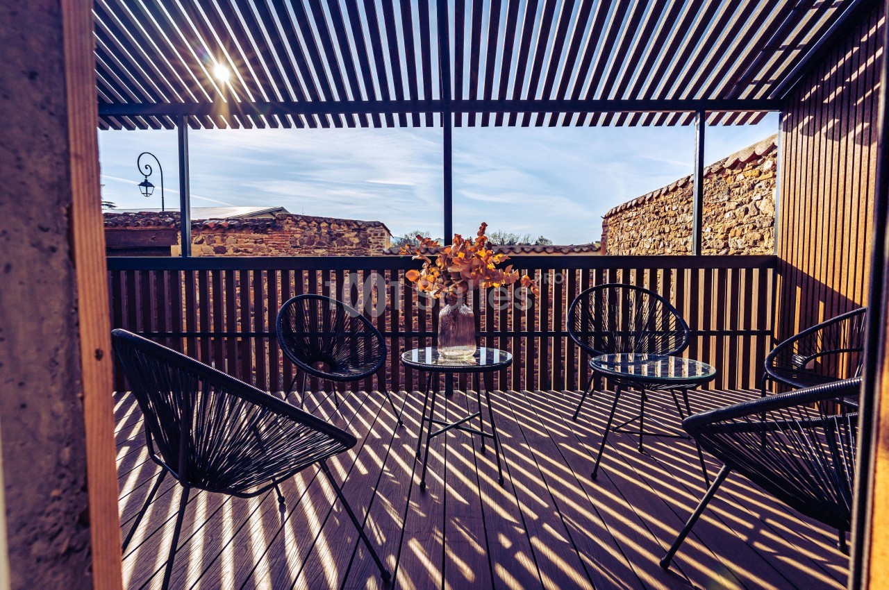 Terrasse en bois avec des fauteuils noirs, une table basse et un vase de fleurs, ombragée par une pergola à lames.