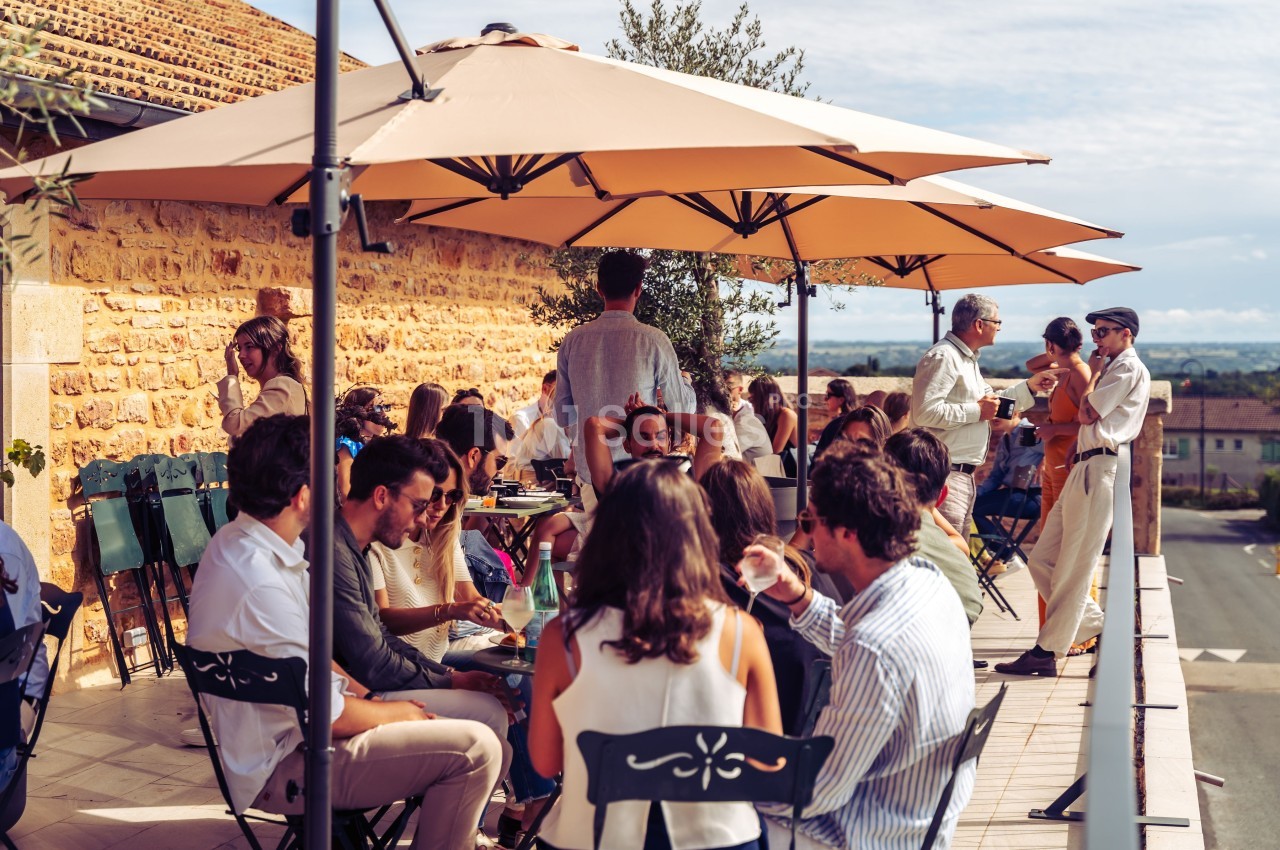 Groupe de personnes assises et debout sur une terrasse ensoleillée, discutant sous de grands parasols beiges.