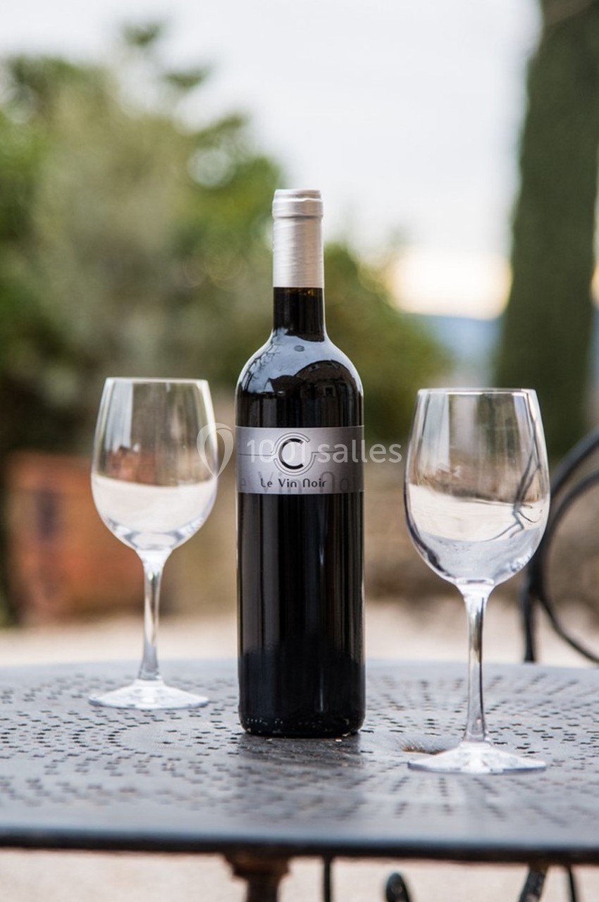 Bouteille de vin rouge posée sur une table en métal avec deux verres à pied, en extérieur.